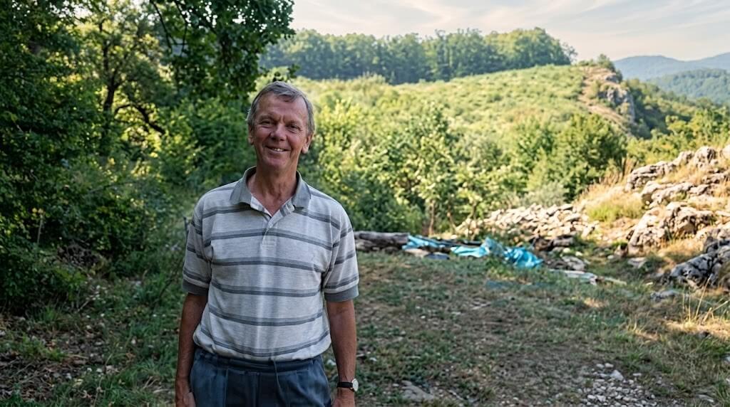 Smiling older man in a striped polo shirt standing outdoors on a dirt path with trees and hills in the background