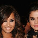 Demi Lovato and Selena Gomez smiling at a formal event with dark background.