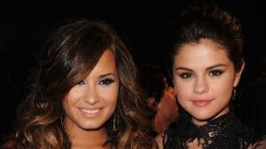 Demi Lovato and Selena Gomez smiling at a formal event with dark background.