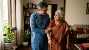 Middle-aged man in blue kurta supporting elderly woman in brown saree with walking stick at home.