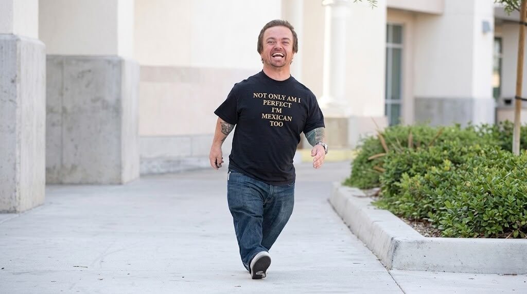Man smiling and walking outdoors wearing a black shirt that says "Not only am I perfect I'm Mexican too"