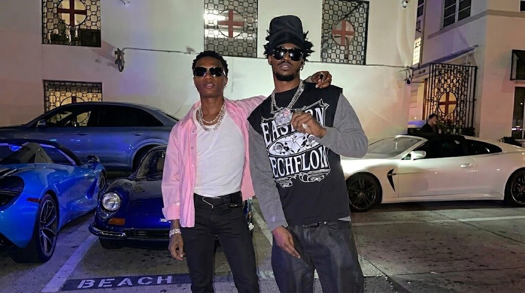Two men wearing sunglasses posing in front of luxury cars in a nighttime urban setting