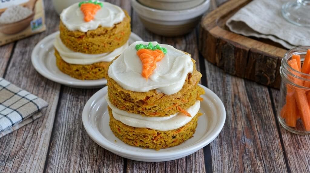 Two stacked carrot cakes with white frosting and a small carrot decoration on top on white plates.