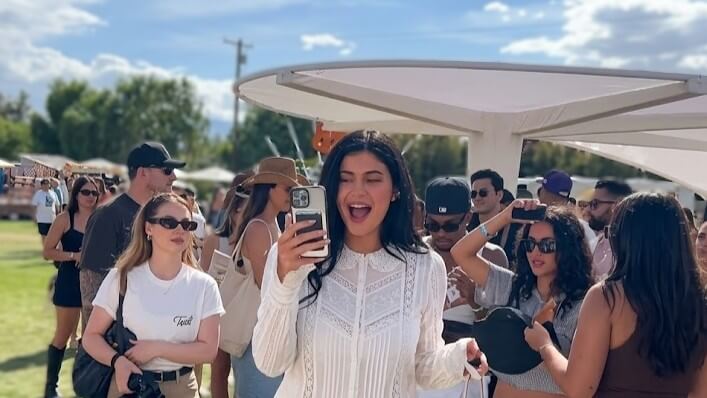 Woman in white lace top smiling and taking a selfie at an outdoor event with a crowd in the background