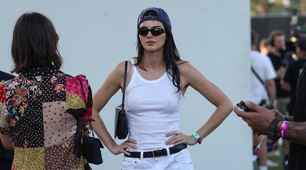 Kendall Jenner wearing a white tank top, white pants, black sunglasses, and a backward blue cap at an outdoor event.