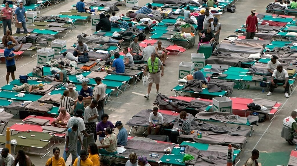People resting and walking among rows of cots in a large emergency shelter or disaster relief center.