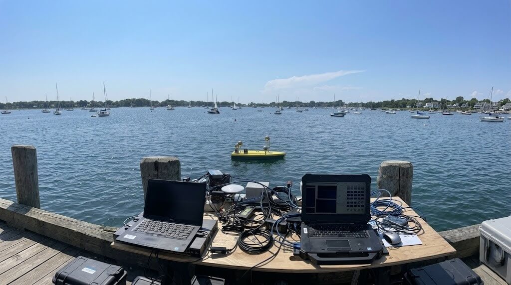 Two laptops and electronic equipment on a dock monitoring a yellow autonomous surface vehicle in a harbor with sailboats.