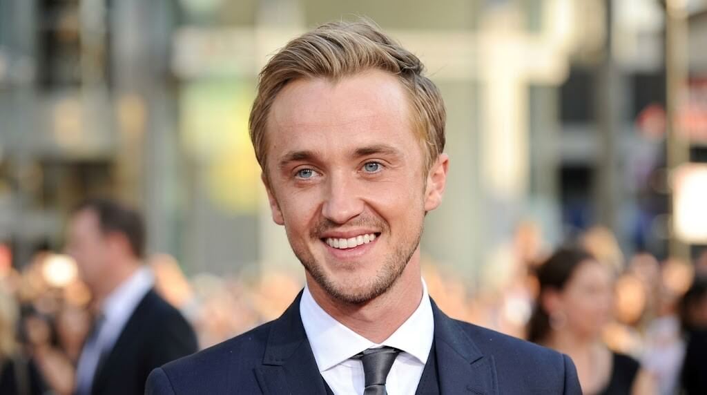 Tom Felton smiling in a dark suit and tie at a public event with blurred crowd background