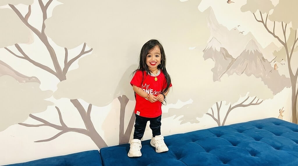 Smiling young girl in a red shirt and black pants standing on a blue couch with a tree and mountain mural background