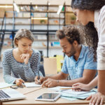 Three young adults collaborating and writing notes at a table with a laptop and tablet in a modern workspace