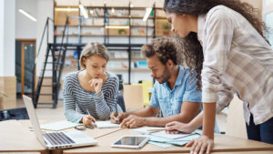 Three young adults collaborating and writing notes at a table with a laptop and tablet in a modern workspace