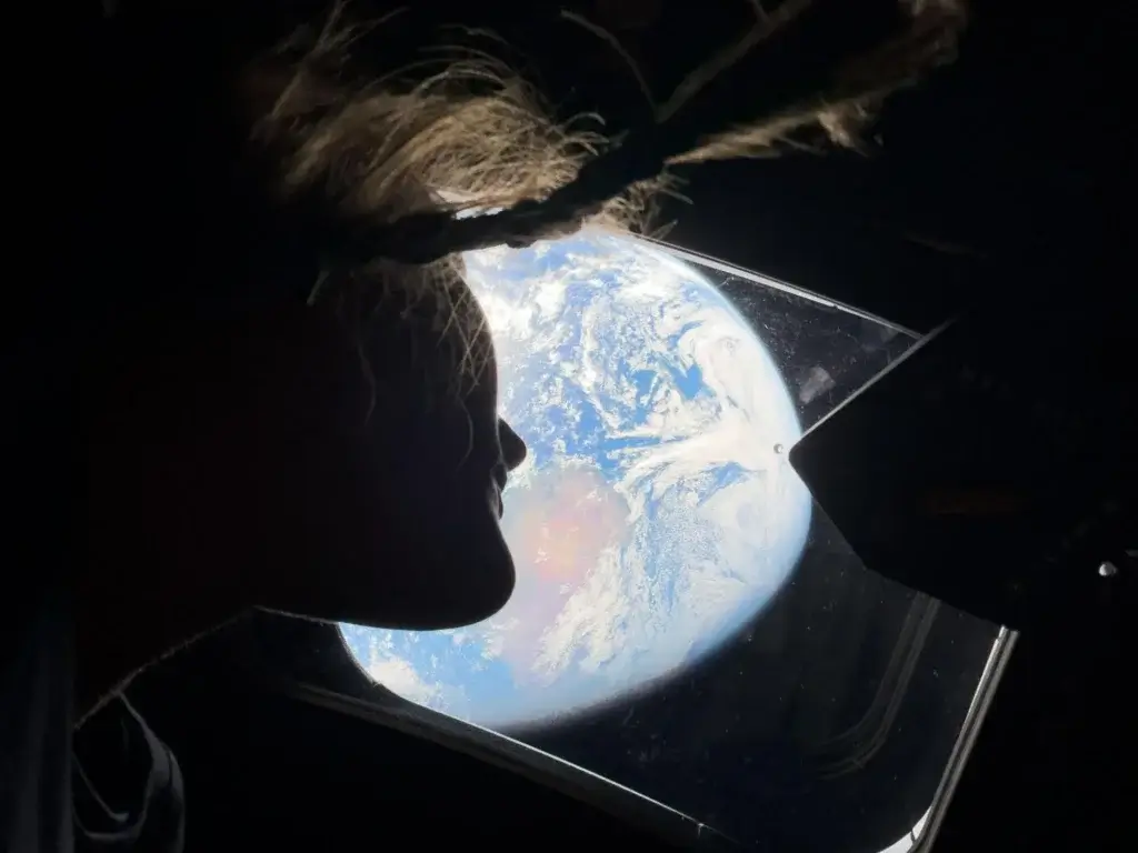 Silhouette of a person with braided hair looking out a spacecraft window at Earth from space.