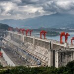 Three Gorges Dam with red cranes on top, surrounded by water and mountains under a cloudy sky