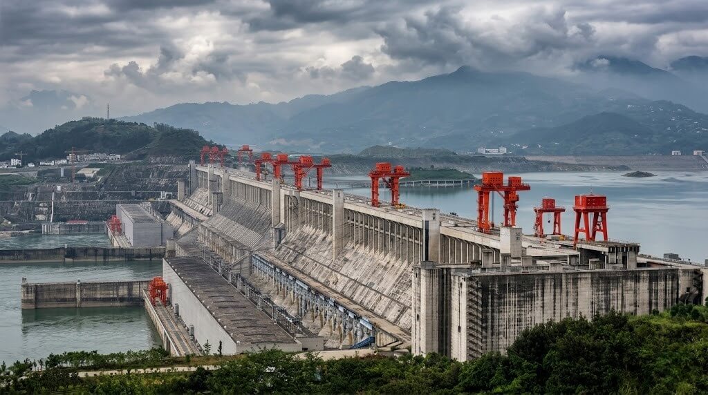 Three Gorges Dam with red cranes on top, surrounded by water and mountains under a cloudy sky