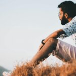 Man with glasses and beard sitting on dry grass wearing a patterned shirt and shorts looking into the distance