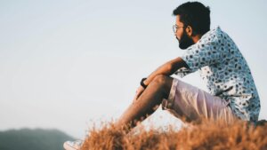Man with glasses and beard sitting on dry grass wearing a patterned shirt and shorts looking into the distance