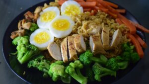 Plate with sliced grilled chicken, hard-boiled eggs, broccoli, carrots, mushrooms, and noodles