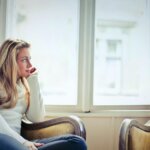 Young woman in a white sweater sitting on a vintage armchair looking out a bright window