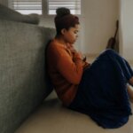 Woman in orange sweater and blue skirt sitting on floor leaning against couch, holding chest with eyes closed