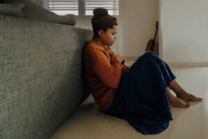 Woman in orange sweater and blue skirt sitting on floor leaning against couch, holding chest with eyes closed