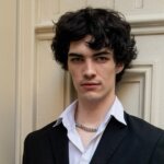 Young man with curly black hair wearing a black blazer, white shirt, and silver chain necklace against a beige wall