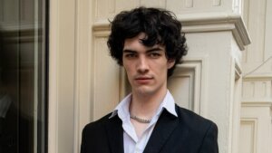 Young man with curly black hair wearing a black blazer, white shirt, and silver chain necklace against a beige wall