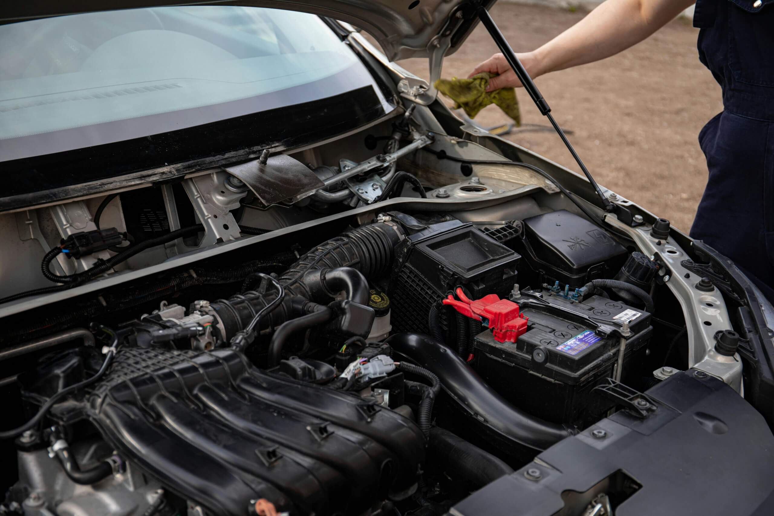 Person cleaning the engine bay of a car with the hood open and a visible car battery with red terminals