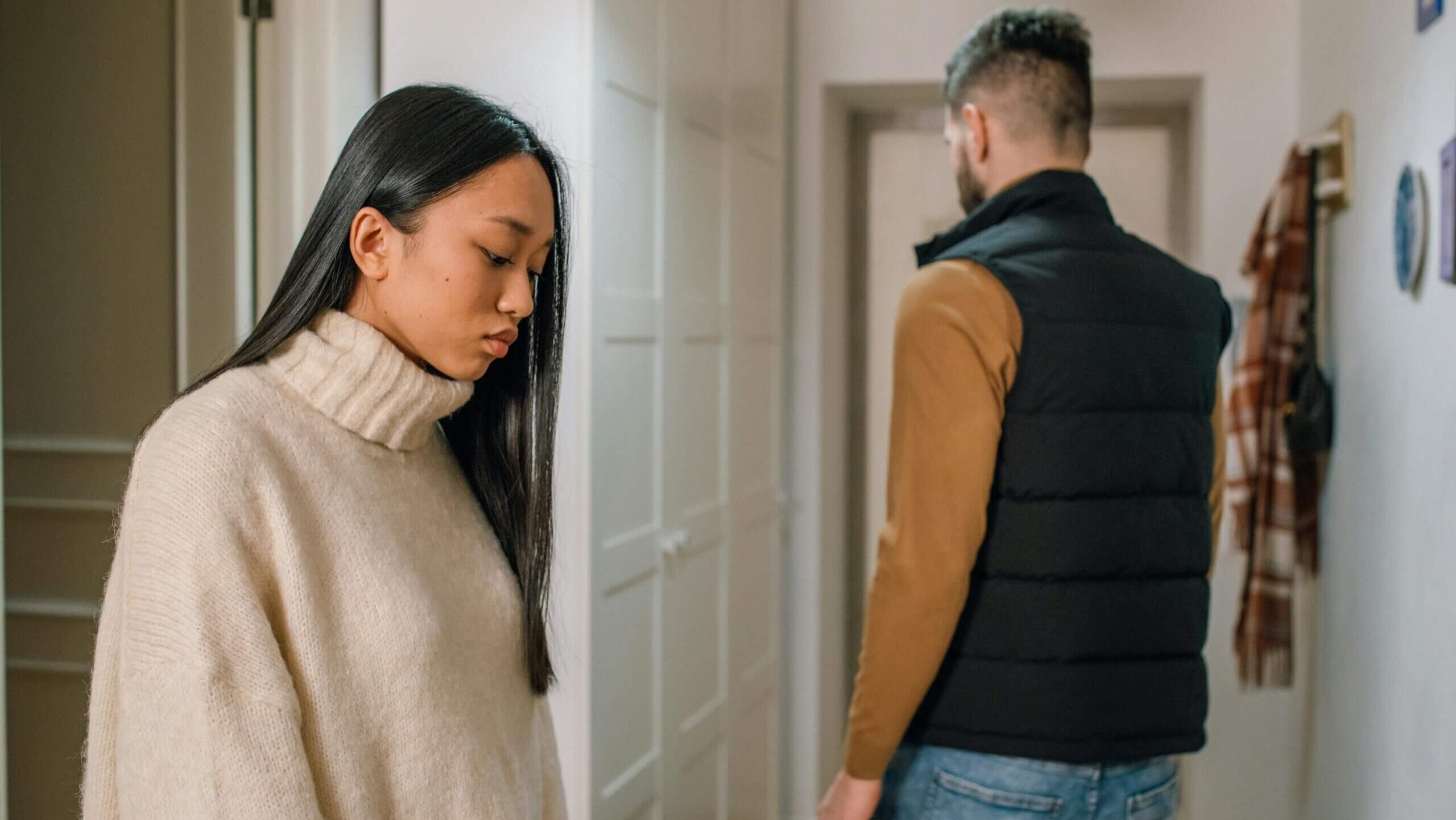 Young woman in beige sweater looking down while man in black vest and jeans walks away in hallway