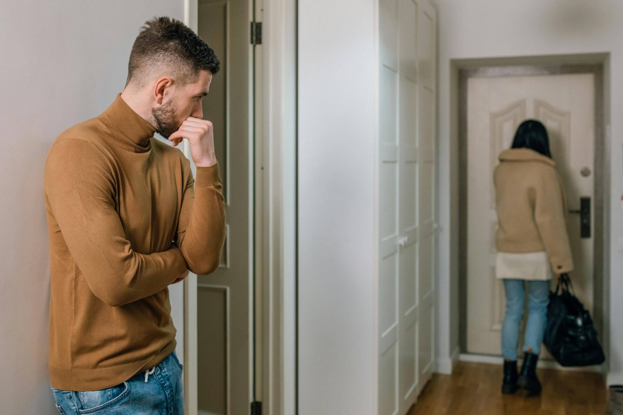 Man in brown turtleneck looking concerned as woman in beige coat and jeans leaves through a door carrying a black bag