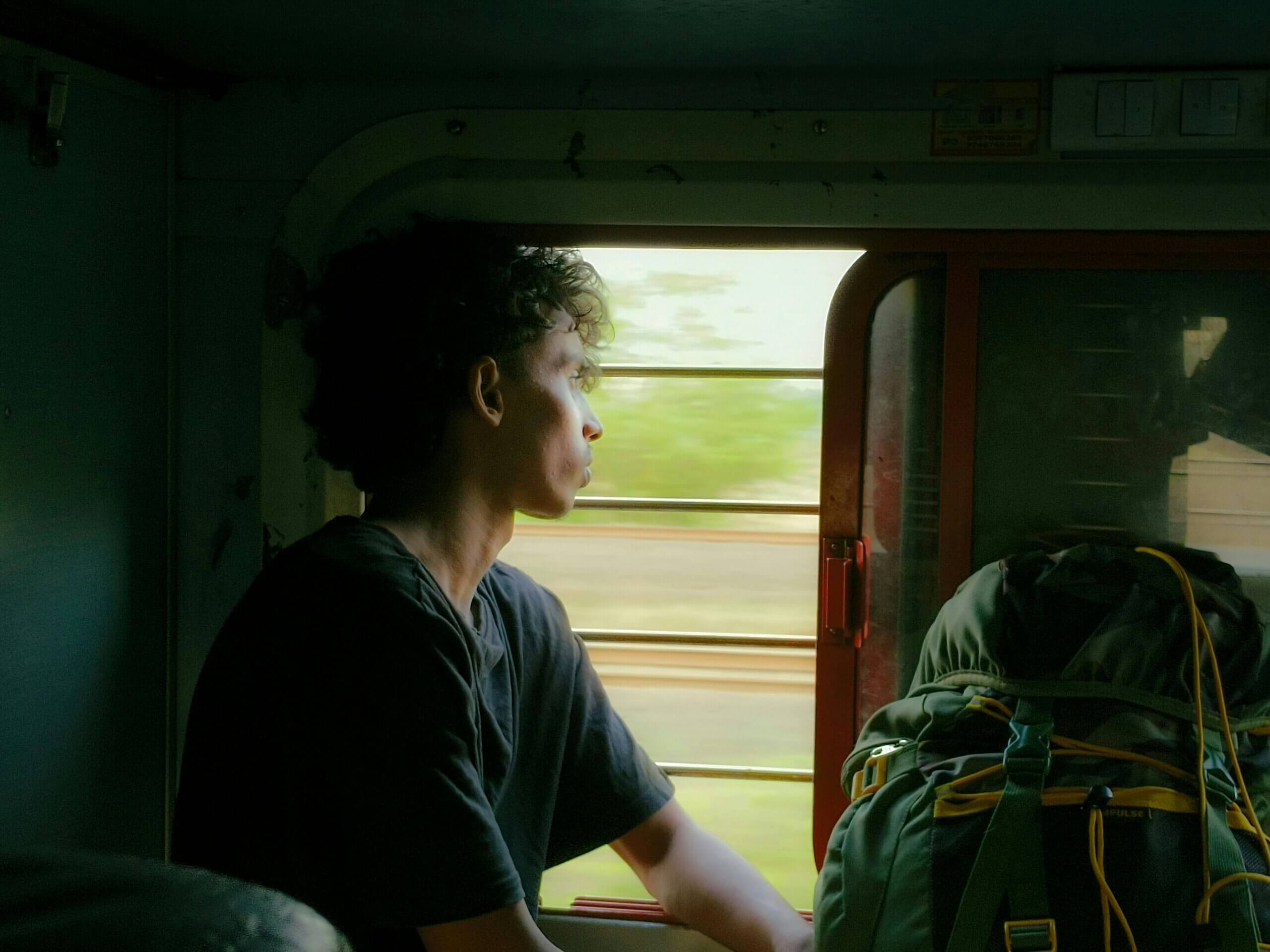 Young man with curly hair looking out a train window next to a large green backpack