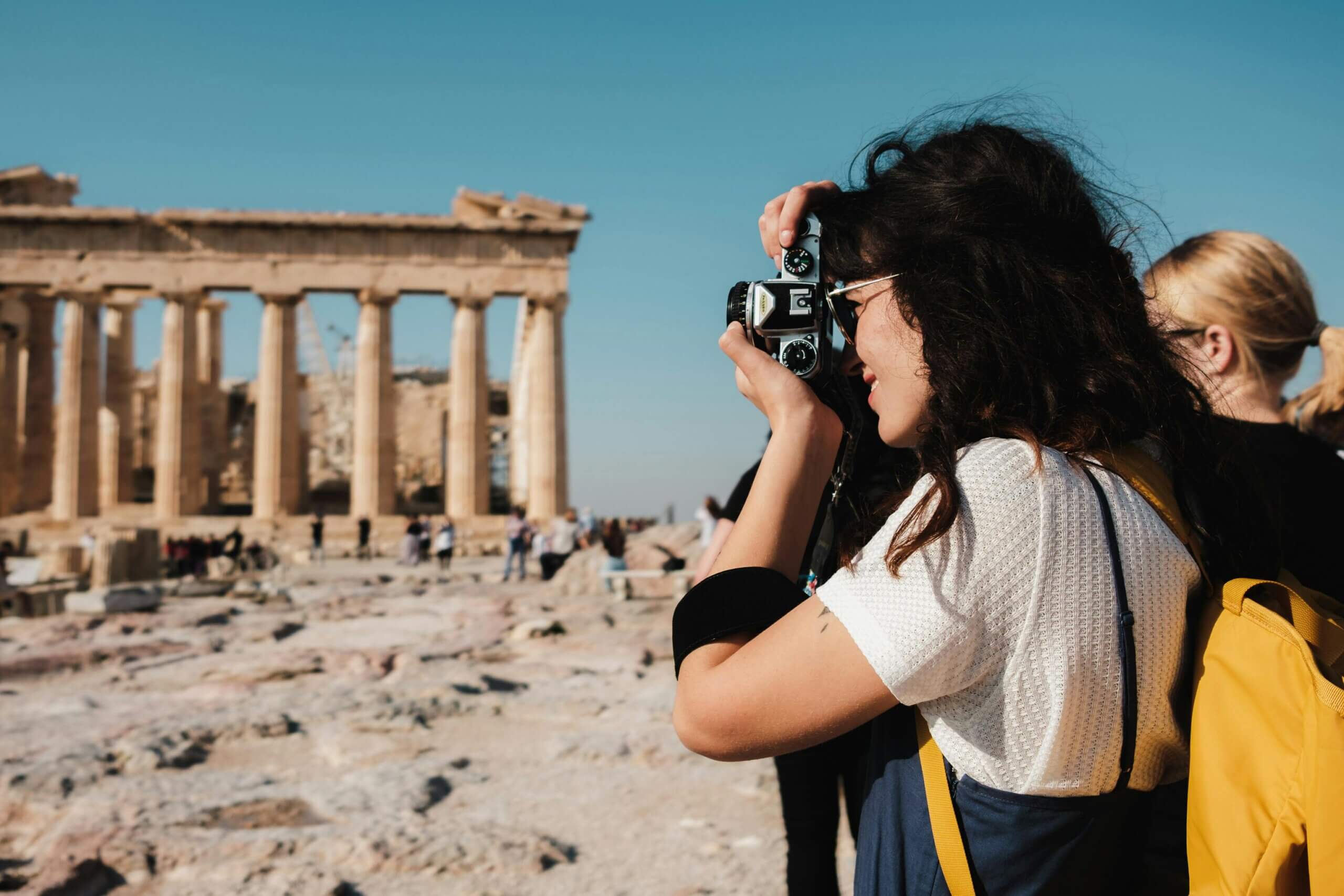 Woman with curly hair and yellow backpack taking a photo of the Parthenon in Athens with a vintage camera