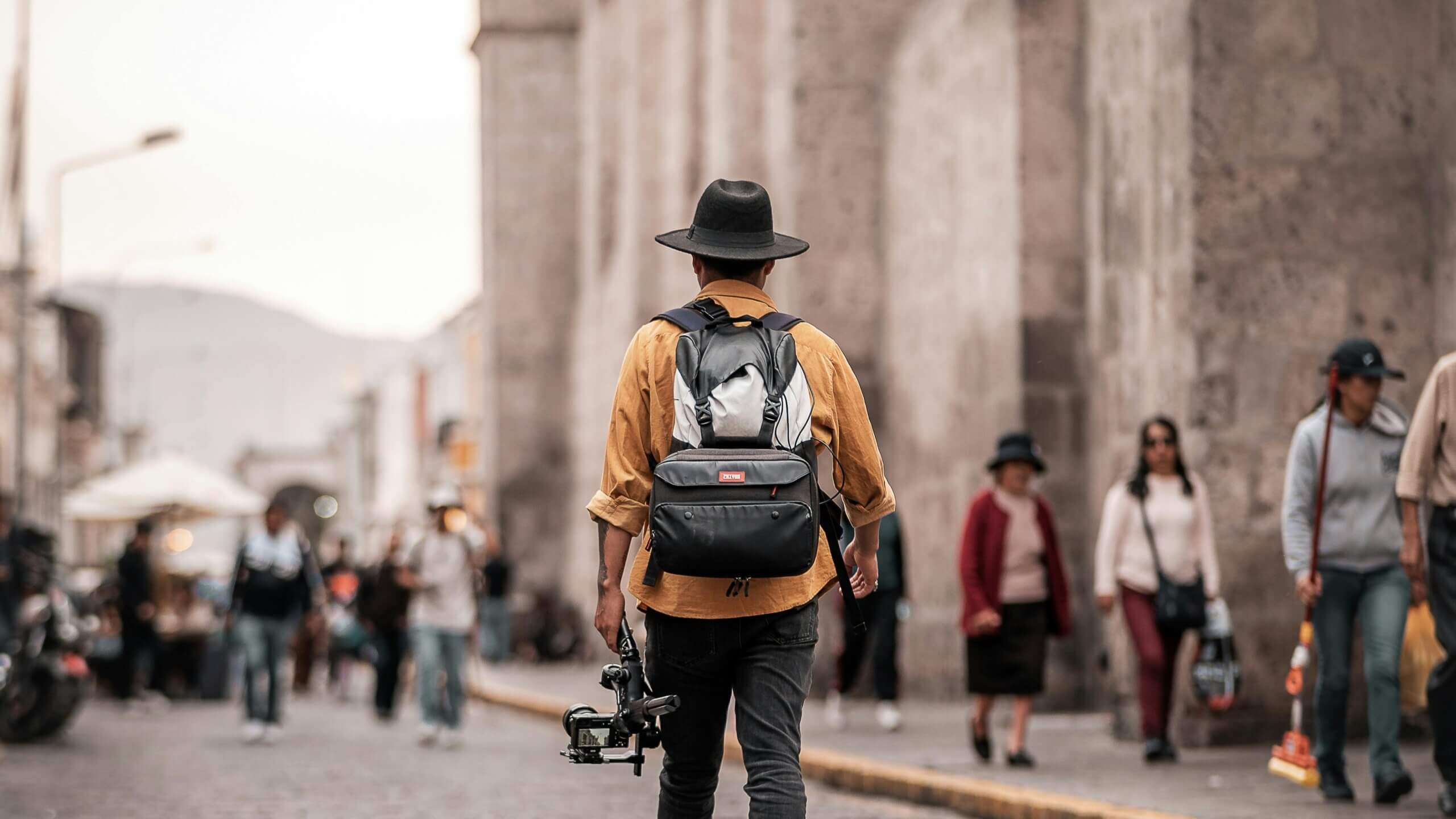 Man wearing a black hat and yellow shirt carrying a camera stabilizer and a black backpack walking on a city street