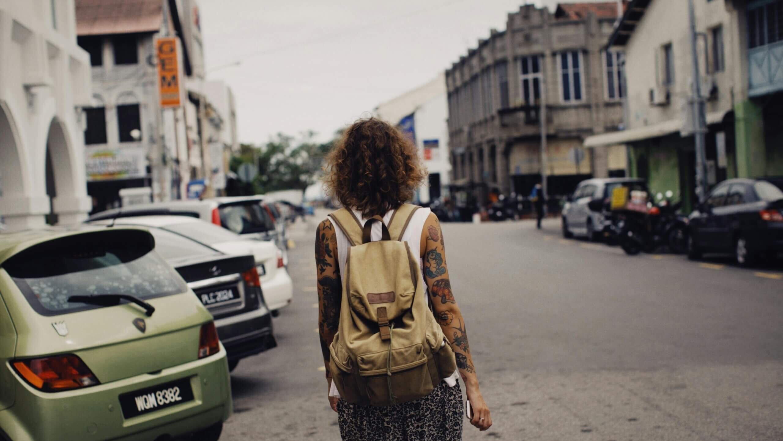 Person with curly hair and tattoos wearing a beige backpack walking down a city street lined with parked cars.