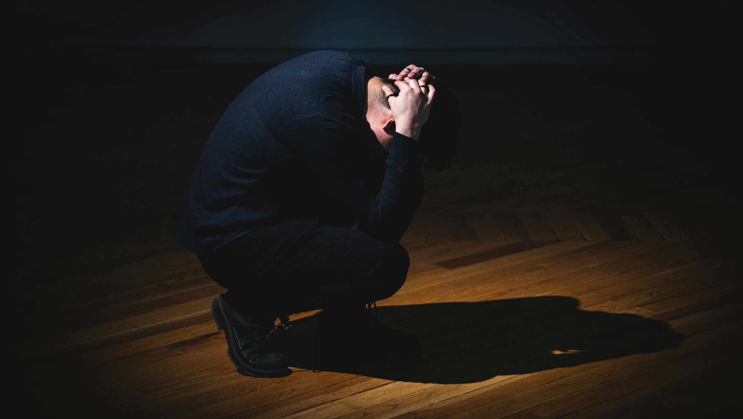 Man crouching on wooden floor with hands clasped on head in dim light