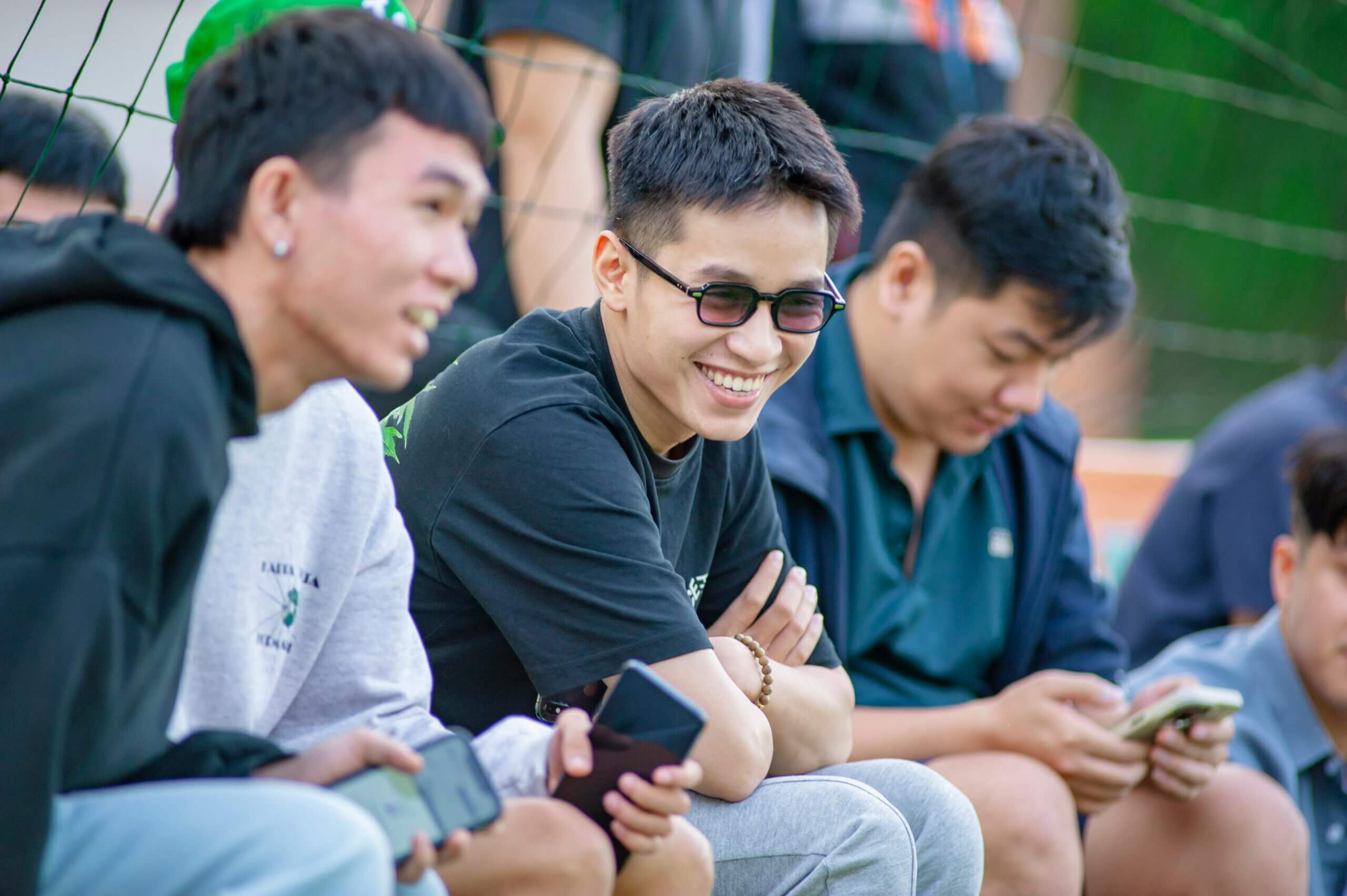 Group of young men sitting outdoors, smiling and using smartphones near a sports net.