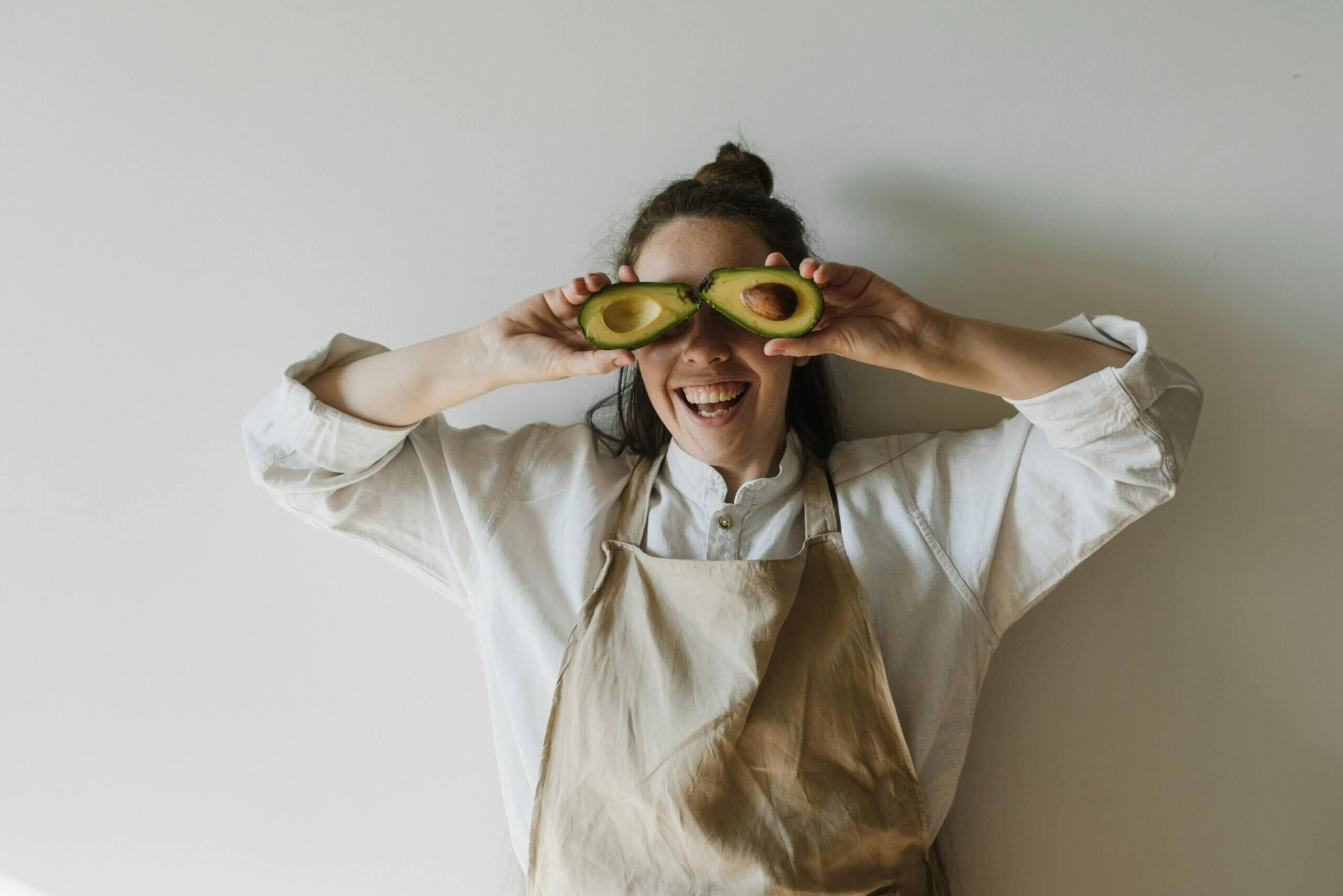 Smiling woman in beige apron holding avocado halves over her eyes against a plain wall background