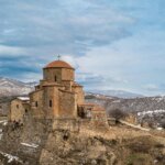 An ancient stone church with a cross on top, situated on a hill with snow-dusted mountains in the background.