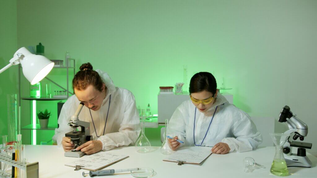 Two scientists in white lab coats working with a microscope and taking notes in a green-lit laboratory.