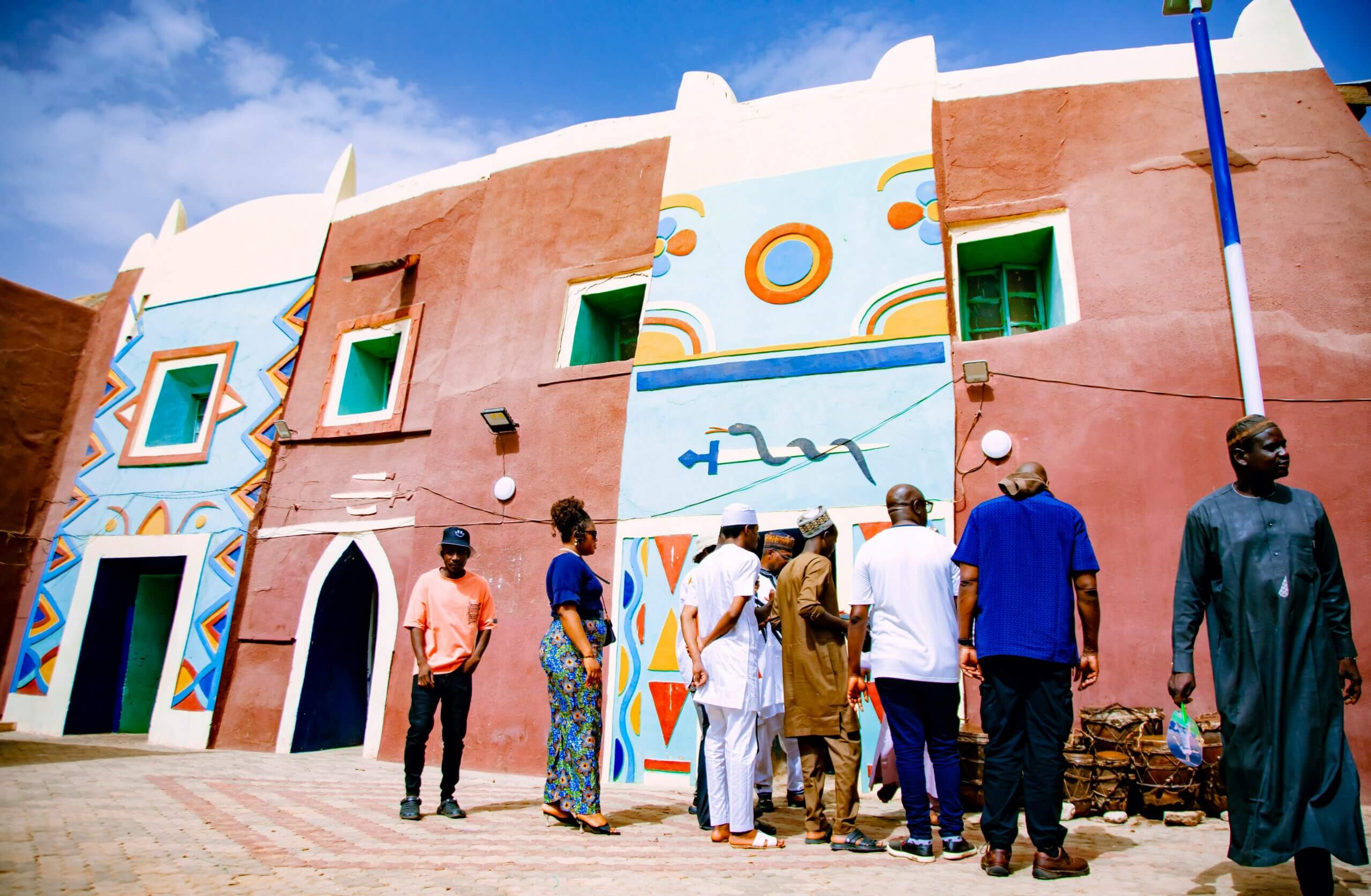 Group of people standing outside a colorful traditional building with geometric and snake mural designs under a clear blue sky