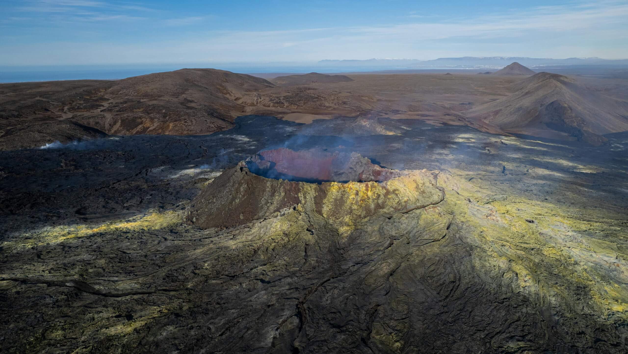 Aerial view of a volcanic crater with smoke and yellow sulfur deposits in a barren landscape.