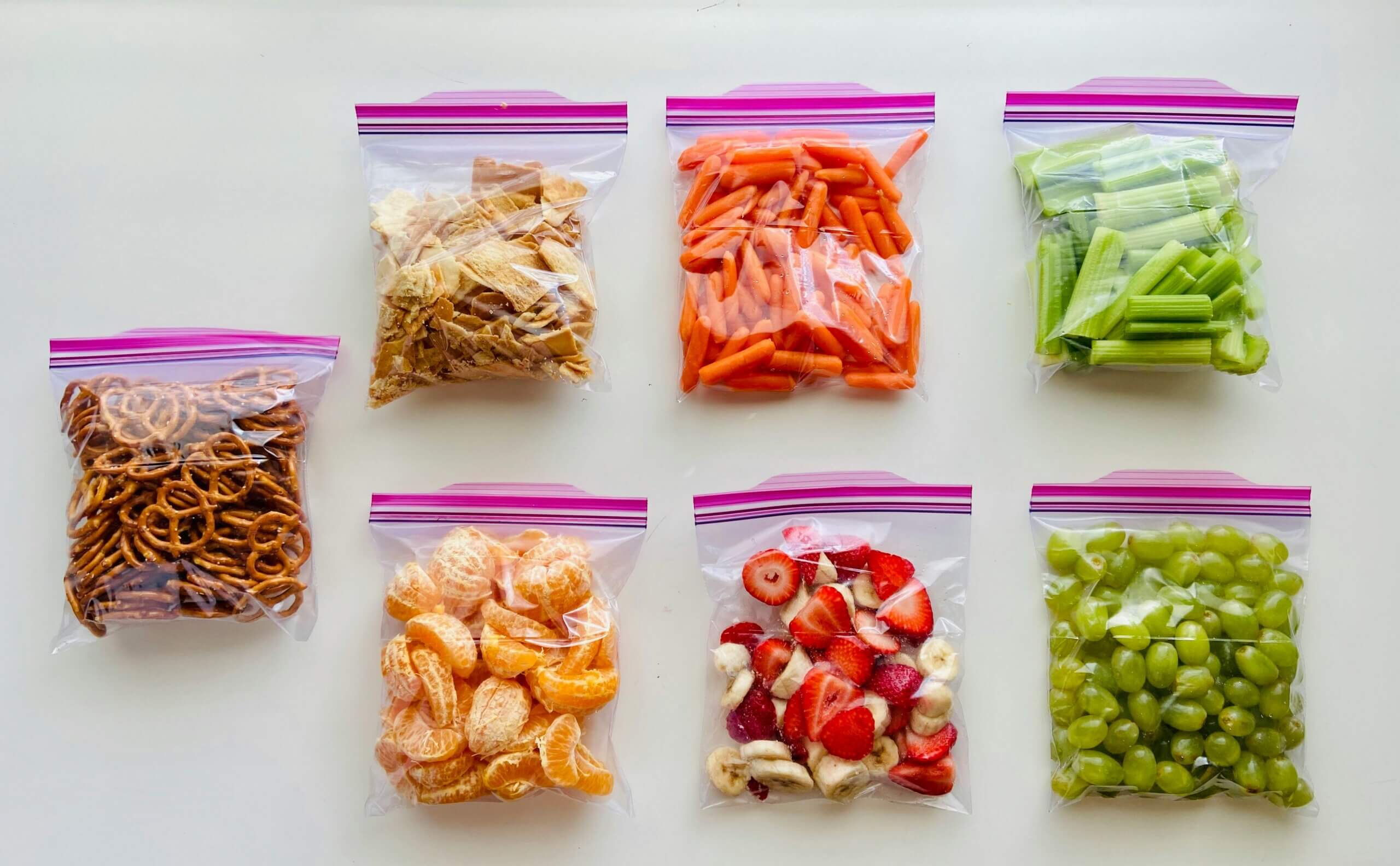 Seven clear plastic bags containing pretzels, crackers, baby carrots, celery, peeled orange segments, sliced strawberries and bananas, and green grapes.