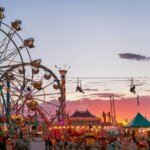 Ferris wheels and zip line at a crowded carnival during sunset with colorful tents and game booths.