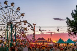 Ferris wheels and zip line at a crowded carnival during sunset with colorful tents and game booths.