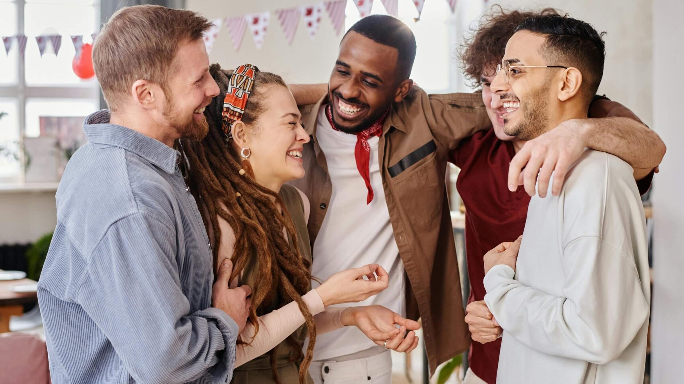 Group of five diverse friends laughing and hugging indoors with festive bunting in the background