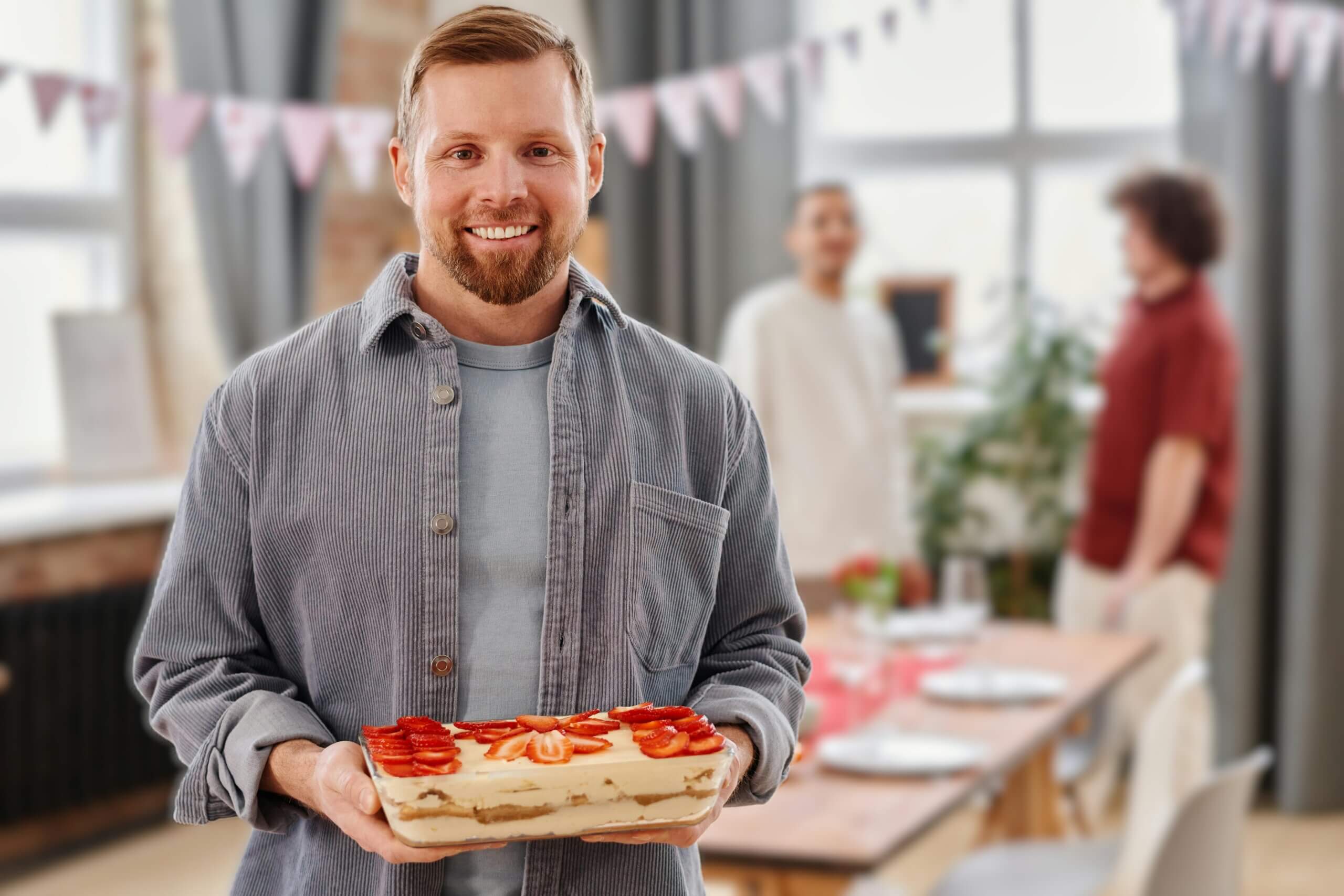 Smiling man holding a strawberry-topped dessert in a decorated room with two people talking in the background.