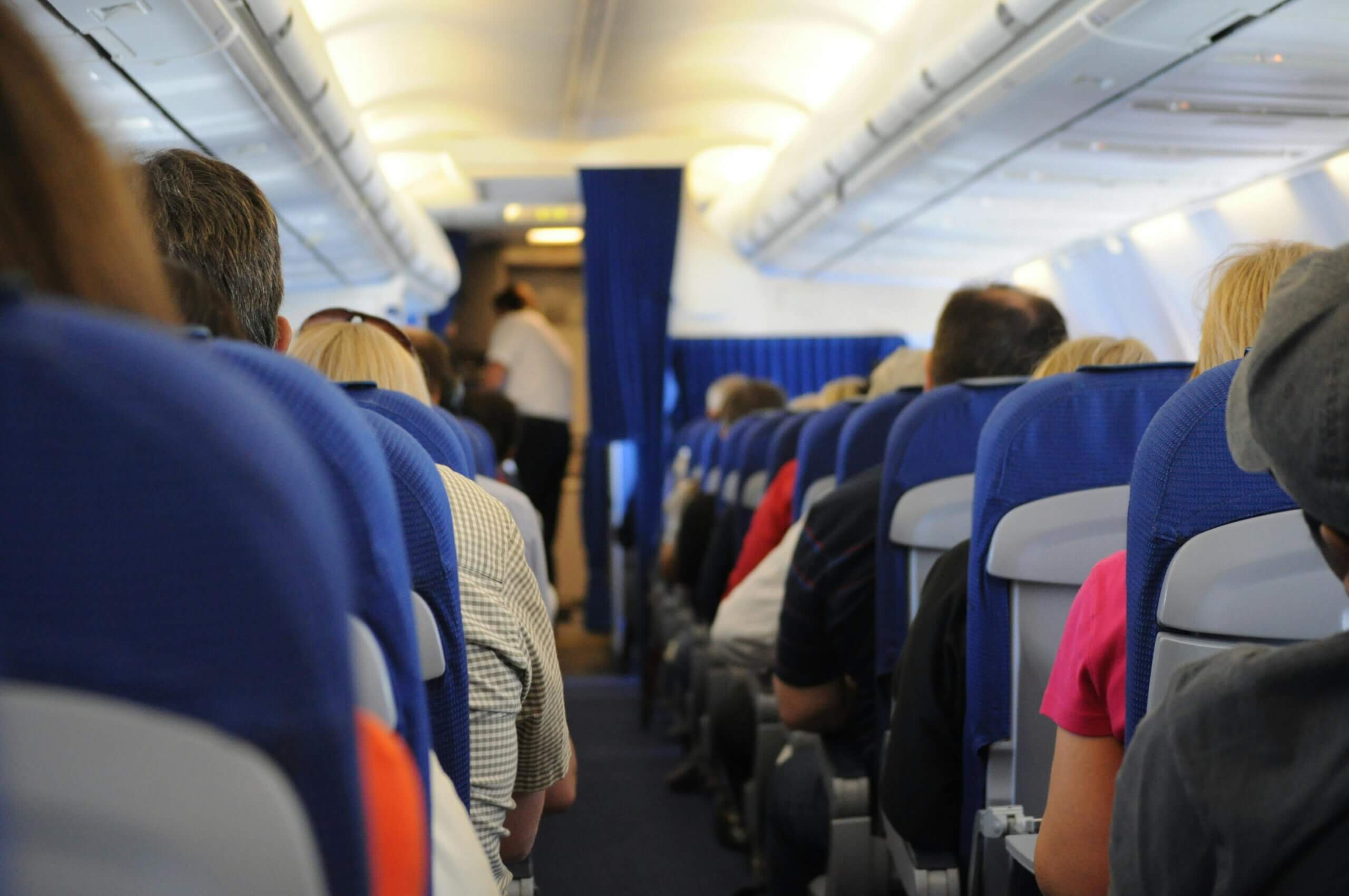 Passengers seated inside an airplane cabin with blue seats and a flight attendant standing near the front.