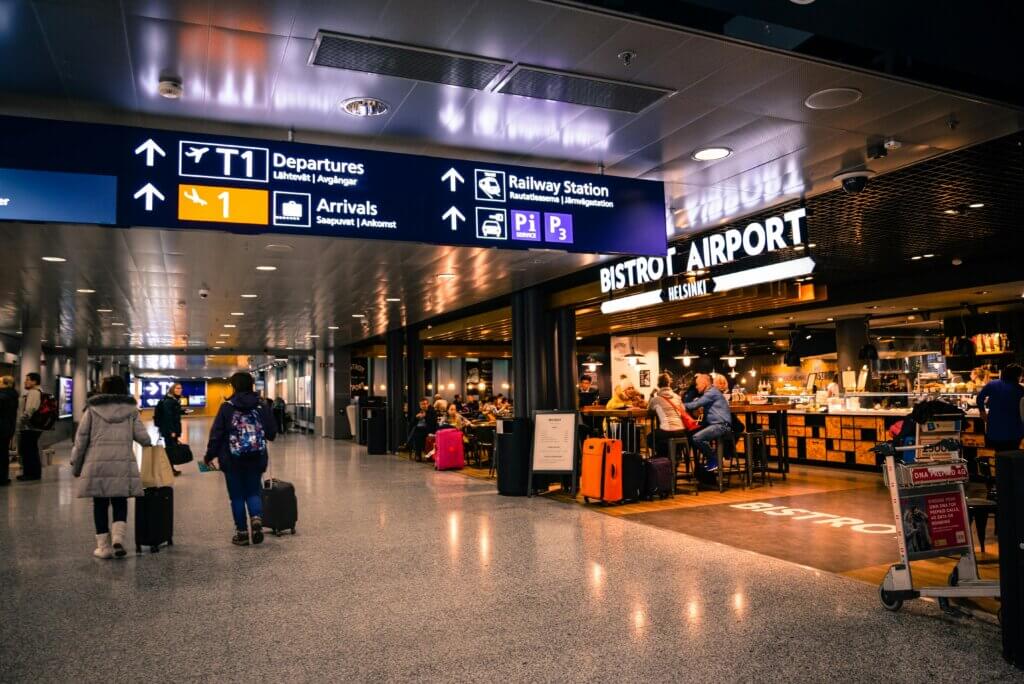 Travelers walk with luggage near Bistrot Airport Helsinki and airport directional signs for departures, arrivals, and railway station.