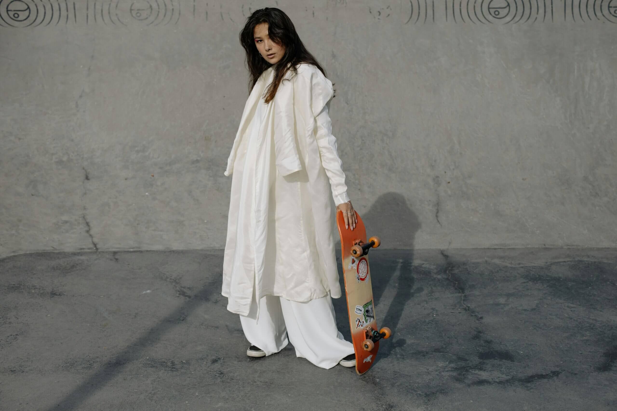 Woman in white outfit holding an orange skateboard with stickers against a concrete wall background