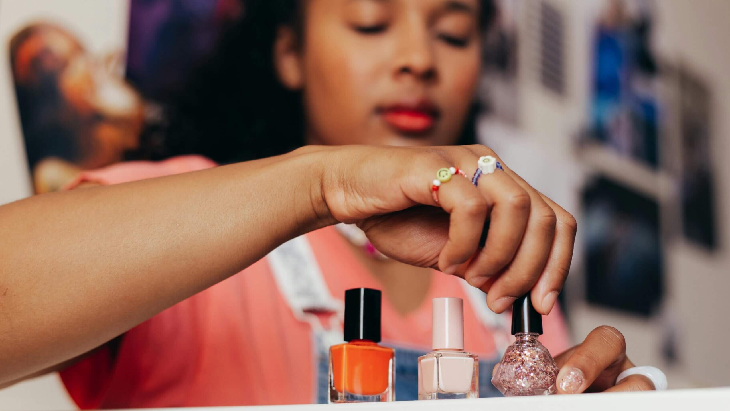 Person selecting a bottle of glittery pink nail polish from three bottles on a white surface.
