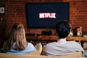 Two people sitting on a couch watching Netflix on a wall-mounted TV in a cozy room with brick walls.